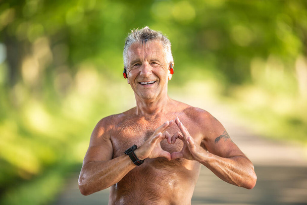Old but fit man with grey hair shows a heart gesture with his fingers as a sign of his healthy cardiovascular system. - Foto, Imagem