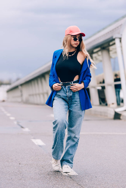 Young sexy blonde hipster woman posing on the street. Wearing blue stylish jacket, jeans and baseball hat and sunglasses. - Foto, afbeelding