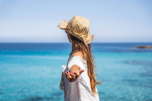 Girl with white dress, brown hat and sunglasses on a paradise beach with crystal turquoise water in the background - Foto, imagen