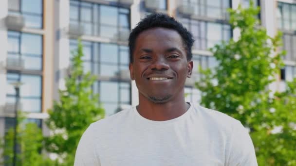Blisko uśmiechnięty afro-amerykański człowiek patrzy aparat fotograficzny na tle nowoczesnego budynku - Materiał filmowy, wideo