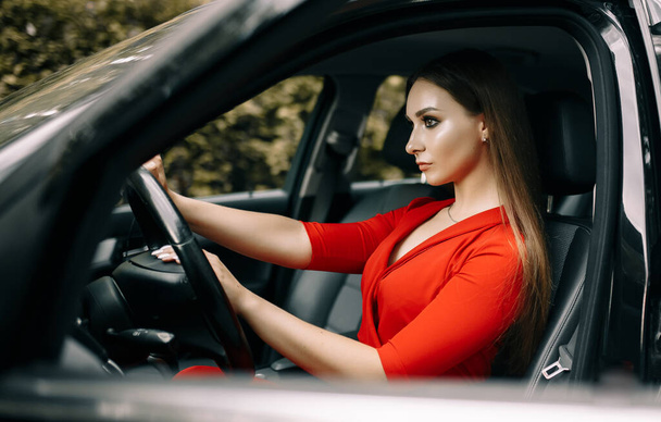 A beautiful young girl in a red overalls sits behind the wheel of a black car on an empty road in the forest - Foto, Bild