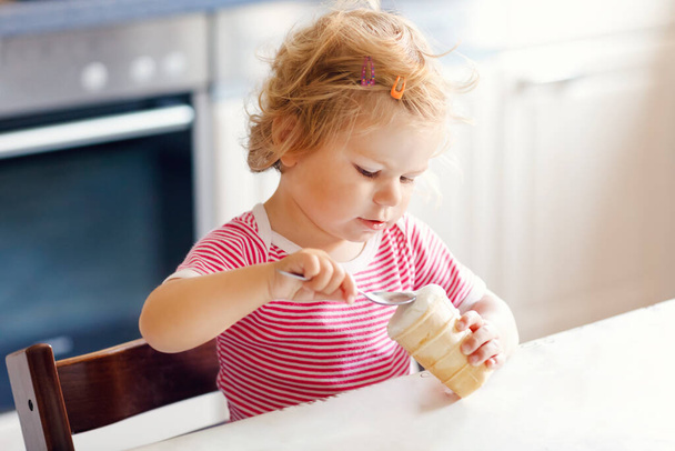 Очаровательная малышка ест из ложки сладкое мороженое в вафельном рожке. питание, ребенок, питание и концепция развития. Симпатичный малыш, дочь с ложкой сидит в кресле и учится есть самостоятельно
 - Фото, изображение