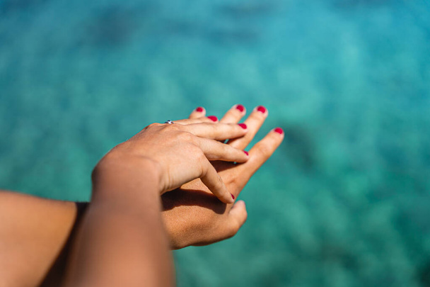 Two girl hands with rings and red nails with turquoise water in the background on a sunny summer day - Fotó, kép