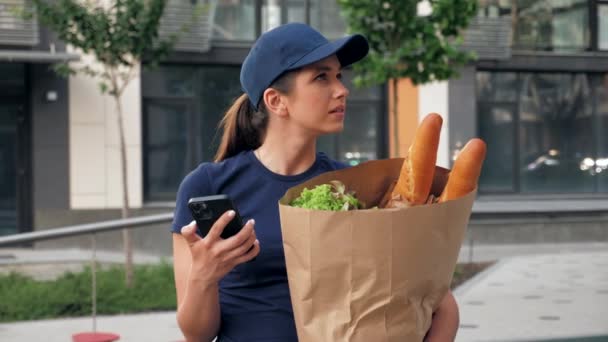 Comida entrega mensajero mujer utiliza teléfono inteligente sostiene bolsa de papel buscando dirección - Metraje, vídeo