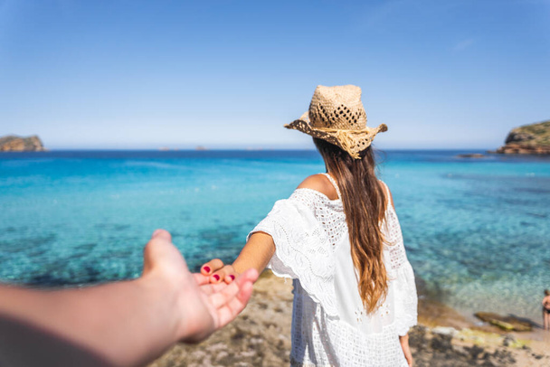 Girl with a white dress and brown hat holding hand with a boy on a paradise beach with crystal turquoise water in the background - Photo, Image