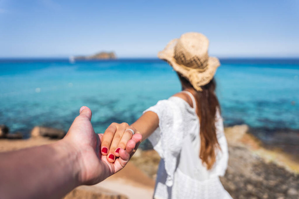 Girl with a white dress and brown hat holding hand with a boy on a paradise beach with crystal turquoise water in the background - Photo, Image