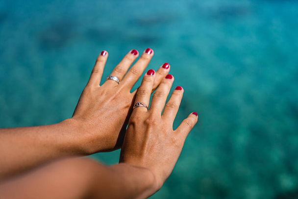 Two girl hands with rings and red nails with turquoise water in the background on a sunny summer day - Photo, Image