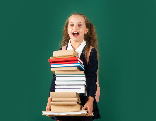 Knowledge day, Smiling funny little schoolkid girl with backpack hold books on green blackboard. Childhood lifestyle concept. Education in school. - Фото, изображение