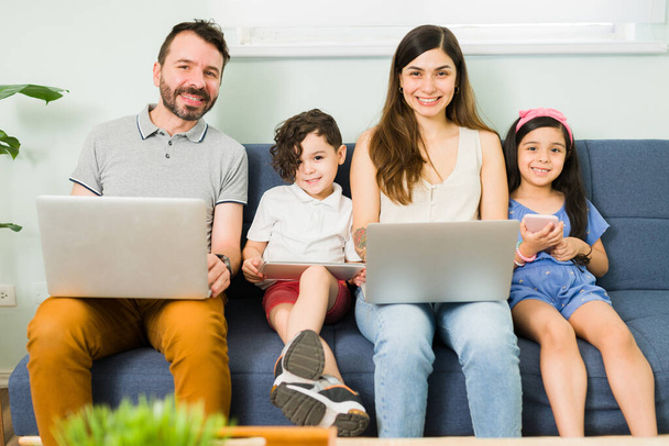 Küçük çocuklarıyla dizüstü bilgisayar tutan gülümseyen bir anne ve Latin babanın portresi oturma odasında elinde bir tablet ve akıllı telefonla oturuyor. - Fotoğraf, Görsel