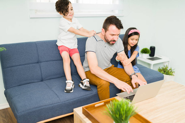 aktive Kinder mit viel Energie, die ihren arbeitenden Vater stören. spanischer Mann versucht, auf dem Laptop zu arbeiten und zu tippen, während seine Kinder versuchen, mit ihm zu spielen - Foto, Bild