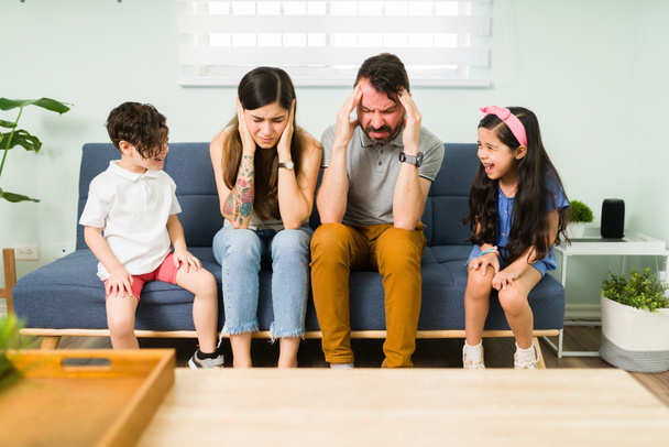 Vihaiset väsyneet vanhemmat peittävät korvansa, kun heidän huonokäytöksiset lapsensa riitelevät. Pikkusisarukset huutaa ja tunne vihainen - Valokuva, kuva