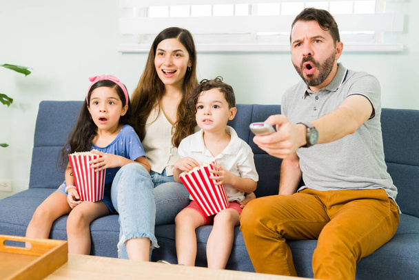 What a shock. Hispanic mom, dad and little children enjoying a family movie on the tv and looking surprised while eating popcorn and snacks - Fotografie, Obrázek