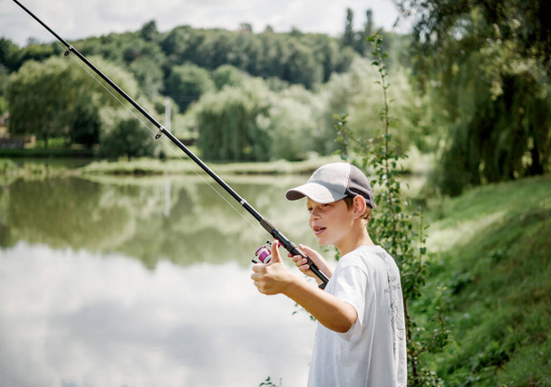 Ένα αγόρι που κρατά καλάμι ψαρέματος και ψαρεύει στη λίμνη, καλοκαιρινές δραστηριότητες και χόμπι για τα παιδιά - Φωτογραφία, εικόνα