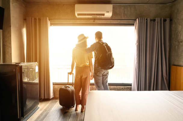 Portrait of young couple tourist standing nearly window, looking to beautiful view outside in hotel/resort bedroom after check-in. Conceptual of couple travel and vacation in their honeymoon period. - Foto, imagen