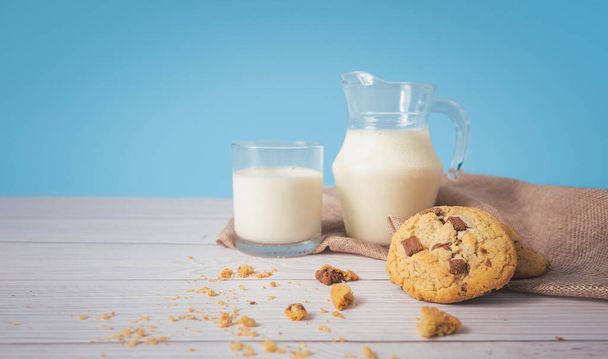 Πεντανόστιμα σπιτικά μπισκότα σοκολάτας, σε συνδυασμό με φρέσκο γάλα σε ποτήρι και κανάτα, τοποθετημένα σε λευκό ξύλινο δάπεδο και μπλε φόντο. Πίνετε το πρωί για υγεία και γερά κόκκαλα. - Φωτογραφία, εικόνα
