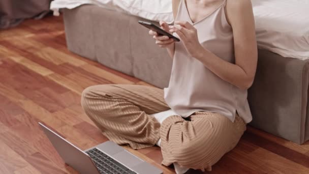 Wysoki kąt nierozpoznawalnej kobiety w piżamie siedzącej na podłodze swojej sypialni, korzystającej z przenośnego komputera i smartfona - Materiał filmowy, wideo