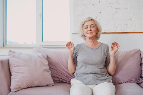 Senior adult smiling woman practicing yoga at home living room. Elderly relaxed female sitting in lotus pose and meditating zen like. - Photo, Image