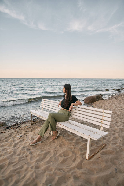 Beautiful brunette woman looking far into the sea horizon. Girls sits on white bench on the beach. Concept of work-life balance and wellbeing. - Foto, immagini