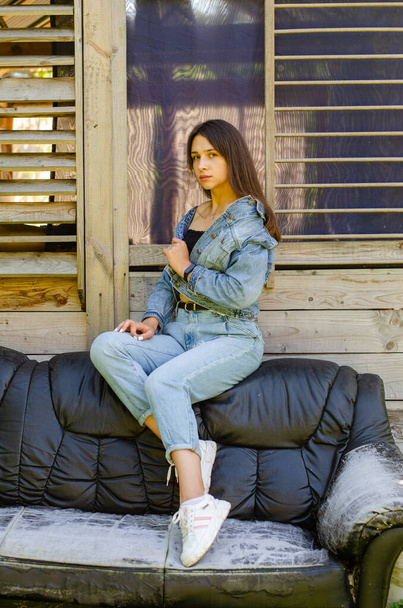 beautiful young girl in jeans clothes posing on leather sofa near house at summer sunny day  - Photo, Image