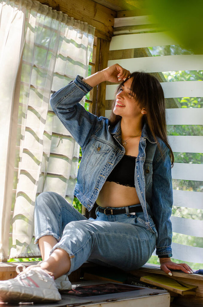 beautiful young girl in jeans clothes posing in wooden house at summer sunny day  - Photo, Image