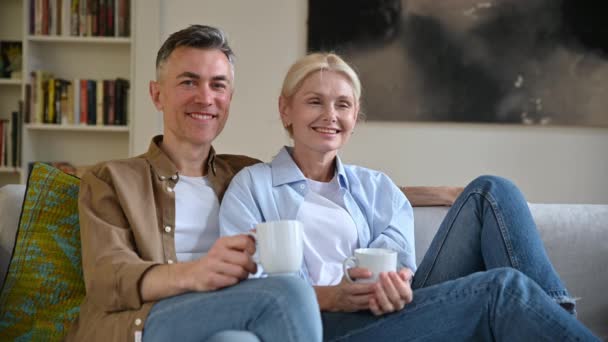 Gelukkig positief volwassen Kaukasisch echtpaar in casual kleding zittend op een comfortabele bank in de woonkamer, theekopjes vasthoudend, kijkend naar de camera, glimlachend. Man en vrouw rusten thuis uit. - Video