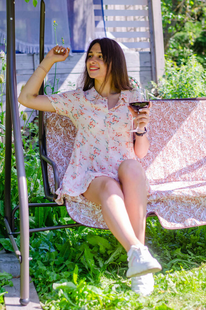 beautiful young girl in summer dress sitting on swing with glass of wine in garden at sunny day - Fotó, kép