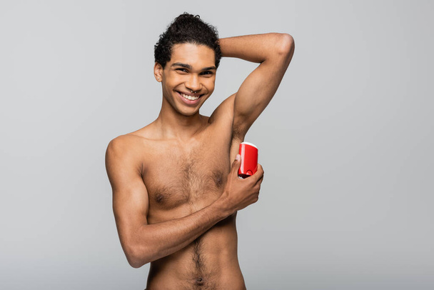 happy african american man looking at camera while applying dry antiperspirant isolated on grey - Foto, imagen