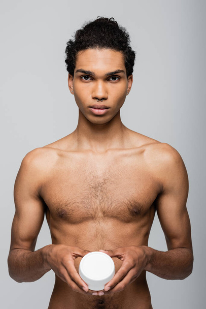 vista frontal do homem americano africano sem camisa segurando creme cosmético isolado em cinza - Foto, Imagem