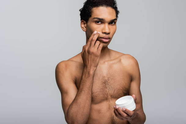 young, shirtless african american man looking at camera and applying face cream isolated on grey - Fotó, kép