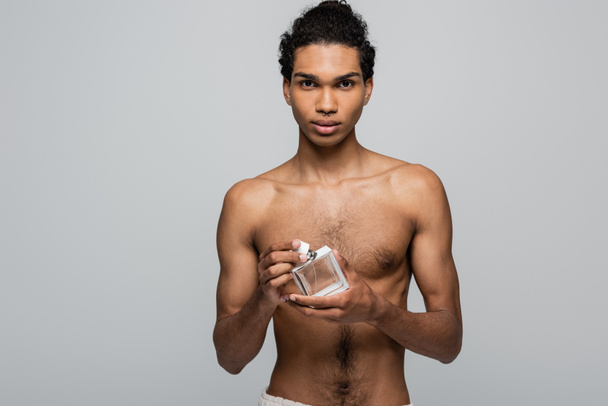 joven, hombre afroamericano sin camisa mirando a la cámara mientras sostiene el perfume aislado en gris - Foto, Imagen