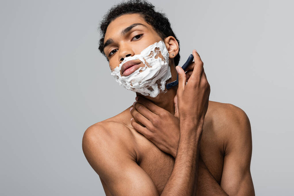 young and shirtless african american man looking at camera while shaving with safety razor isolated on grey - Fotó, kép