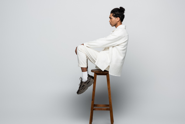 side view of african american man sitting on high stool in white suit and sneakers on grey background - Foto, immagini