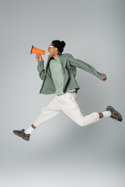 stylish african american guy shouting in megaphone while levitating on grey background - Фото, изображение