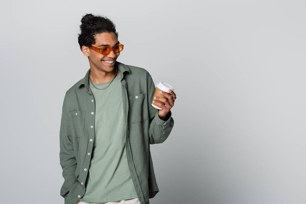 веселый африканский американец в стильных очках, смотрящий на бумажную чашку, изолированную на сером - Фото, изображение