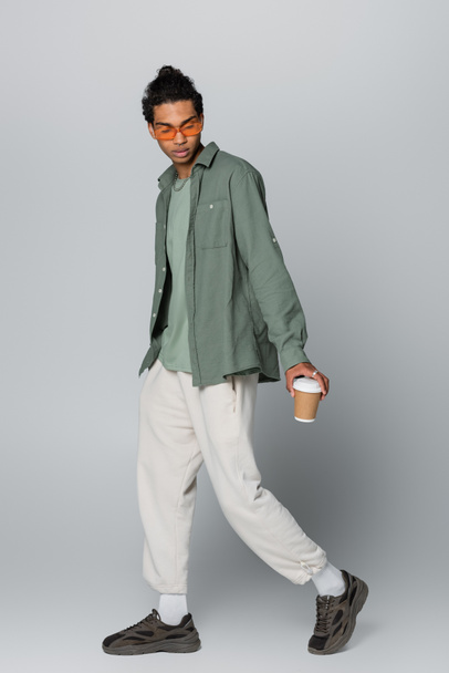 full length view of stylish african american guy walking with coffee to go on grey background - Fotó, kép