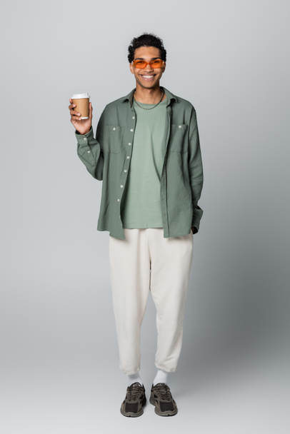 full length view of african american guy in trendy clothes holding takeaway drink isolated on grey - Фото, изображение