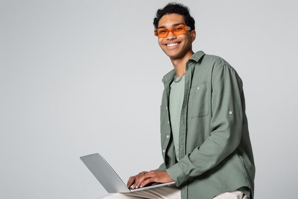 cheerful african american guy in stylish eyeglasses typing on laptop and smiling at camera on grey background - Foto, Imagen