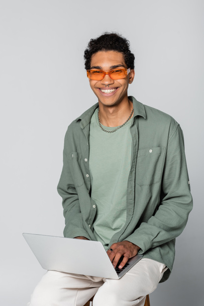 happy african american man in orange eyeglasses looking at camera and typing on laptop on grey background - Fotoğraf, Görsel