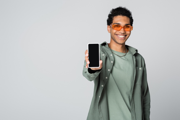 smiling african american man in trendy eyeglasses showing smartphone with blank screen isolated on grey - Photo, Image