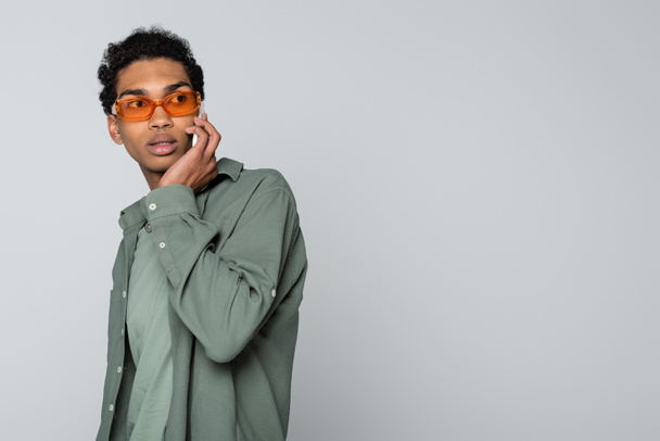 reflexivo afroamericano chico en gafas naranjas hablando en teléfono inteligente aislado en gris - Foto, Imagen