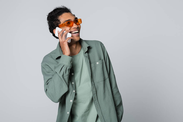 trendy african american man laughing while talking on mobile phone isolated on grey - Photo, Image