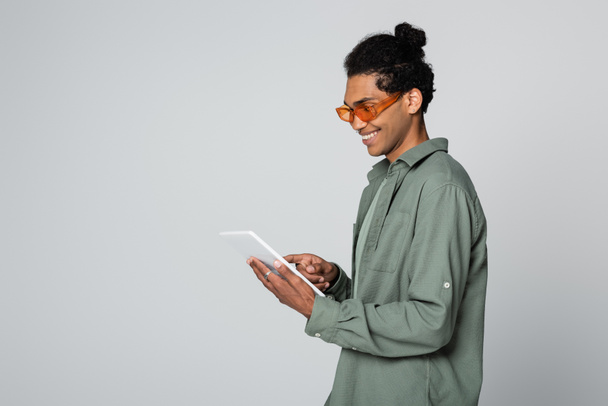 улыбающийся и модный африканский американец, использующий цифровой планшет, изолированный на сером - Фото, изображение
