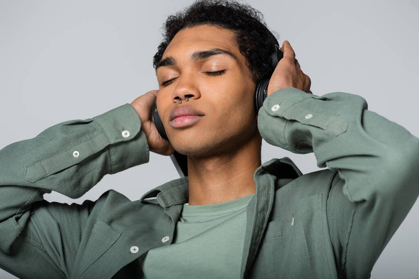 jeune homme afro-américain toucher écouteurs tout en écoutant de la musique avec les yeux fermés isolé sur gris - Photo, image