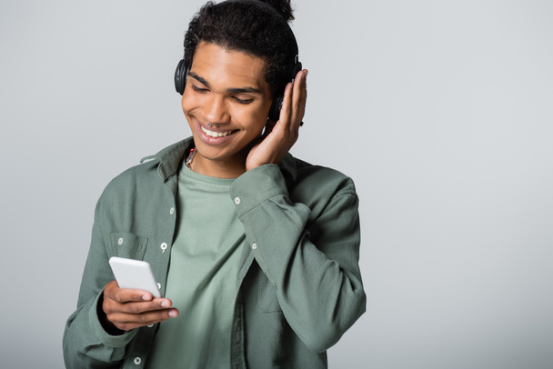 stylish african american guy looking at smartphone while listening music in wireless headphones isolated on grey - Foto, afbeelding