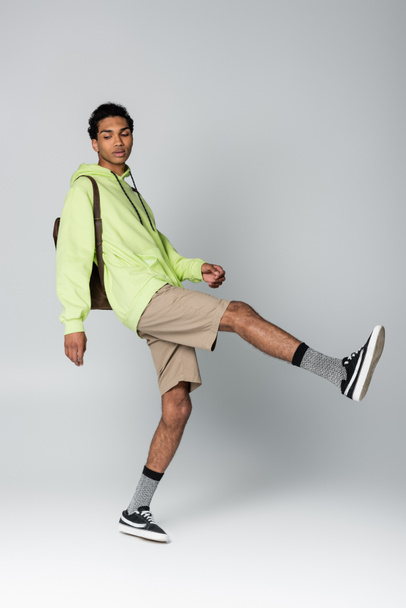 trendy afrikanisch-amerikanischer Mann in Kapuzenpulli und Shorts posiert auf einem Bein isoliert auf grau - Foto, Bild