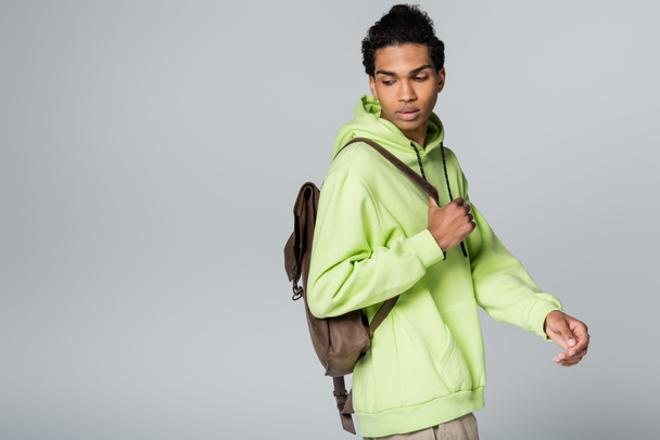 african american guy wearing green hoodie and carrying leather backpack isolated on grey - Foto, imagen