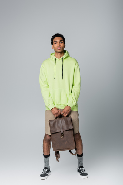 full length view of african american guy in green hoodie standing with leather backpack isolated on grey - 写真・画像
