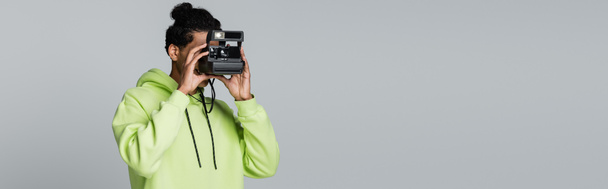 Αφροαμερικάνος με κουκούλα φωτογραφίζει μια παλιά κάμερα σε γκρι λάβαρο. - Φωτογραφία, εικόνα