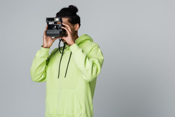 young african american man in green hoodie taking picture on vintage camera isolated on grey - Zdjęcie, obraz