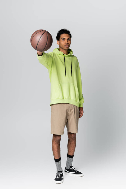 african american guy in green hoodie and beige shorts showing basketball on grey background - 写真・画像